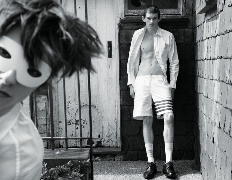 Model Joe Bottomer in an unbuttoned shirt and white shorts standing outside while a masked figure looks in the foreground.