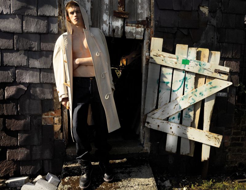 Model Joe Bottomer wearing an open cream hooded coat shirtless with black trousers against a weathered slate wall.