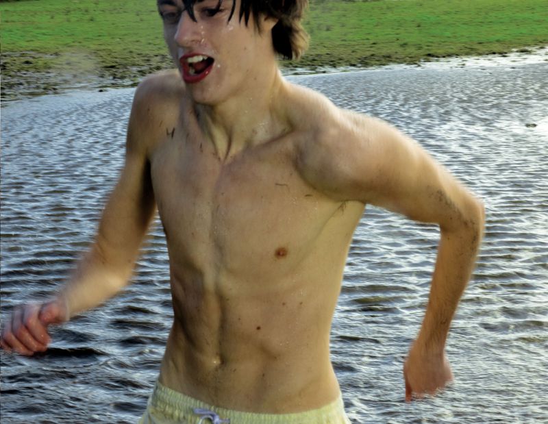 Shirtless young man with wet dark hair running through natural shallow water with a green grassy bank in the background.
