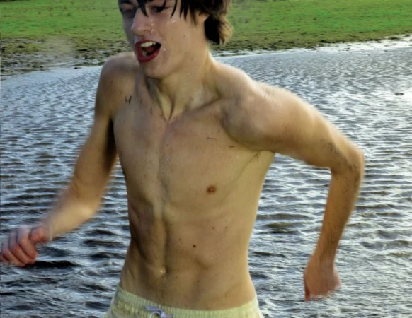 Shirtless young man with wet dark hair running through natural shallow water with a green grassy bank in the background.