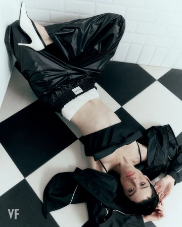 Model Dantè Kedde lying on a black and white checkered floor wearing black track pants and white pointed-toe heels.