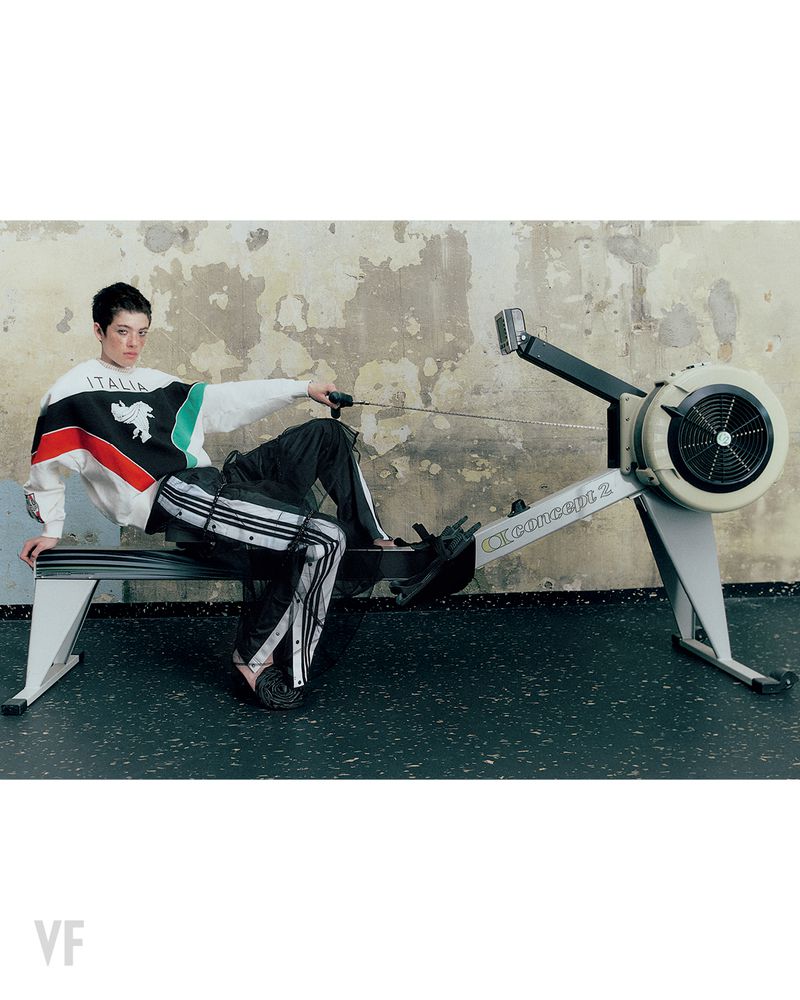 Model posed on an indoor rowing machine wearing an Italia graphic sweatshirt and black track pants against a textured wall.