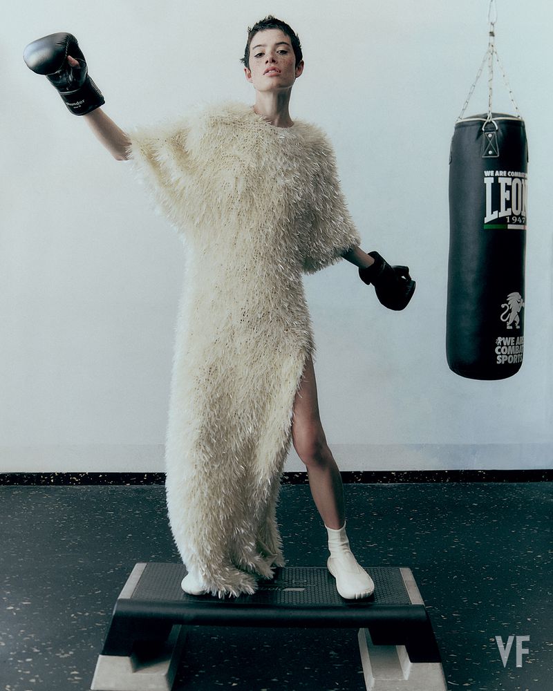 Model Dantè Kedde in a textured cream gown and black boxing gloves standing on an exercise step bench beside a punching bag.