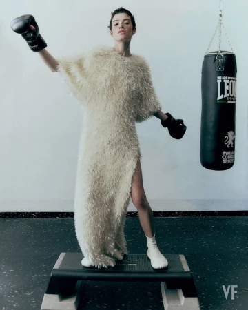 Model Dantè Kedde in a textured cream gown and black boxing gloves standing on an exercise step bench beside a punching bag.
