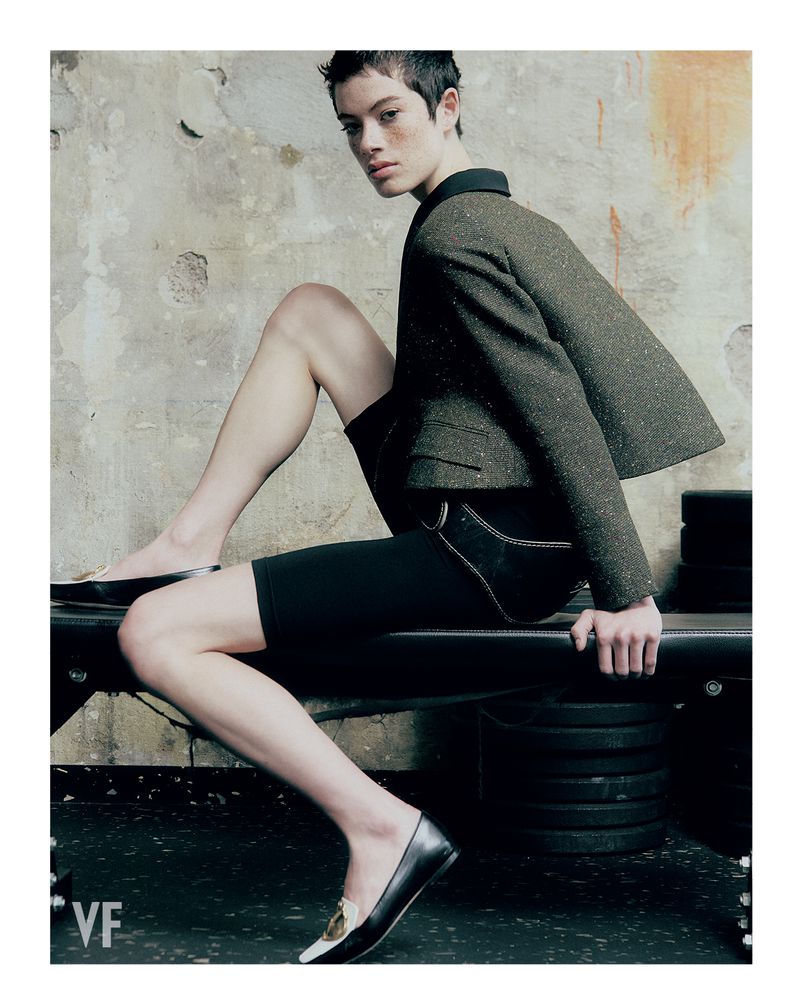 Model with cropped hair seated on a workout bench wearing a structured cropped jacket, black cycling shorts, and black flats.