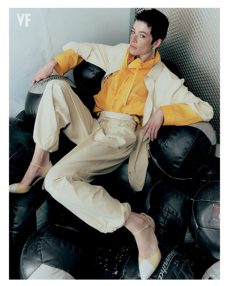 Model Dantè Kedde in a yellow hooded jacket and cream track pants reclining on a pile of black medicine balls.
