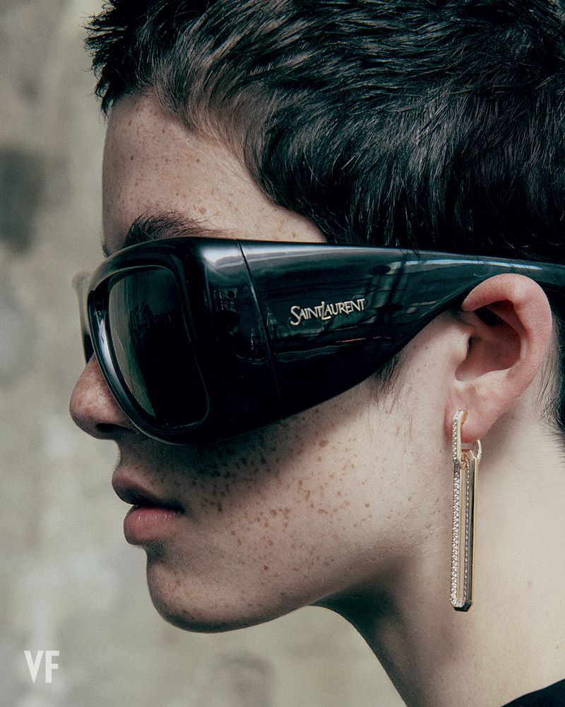 Close-up profile of a model with freckled skin wearing black oversized Saint Laurent sunglasses and a long dangling drop earring.