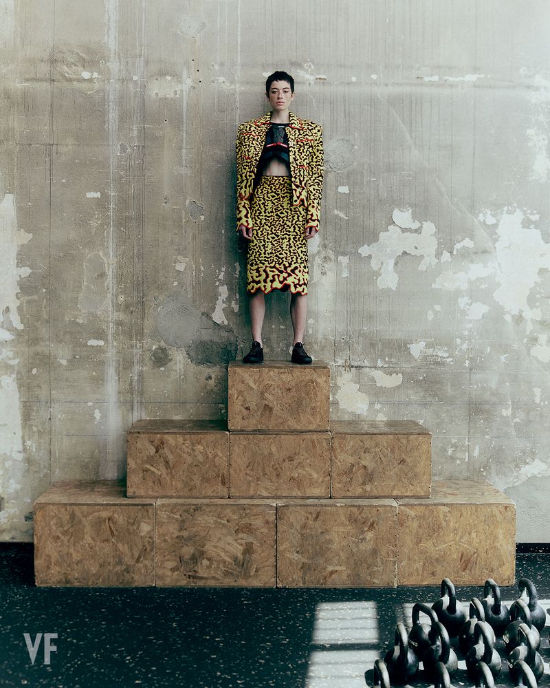 Model Dantè Kedde standing atop stacked wooden boxes in a patterned yellow suit with kettlebells in the foreground.