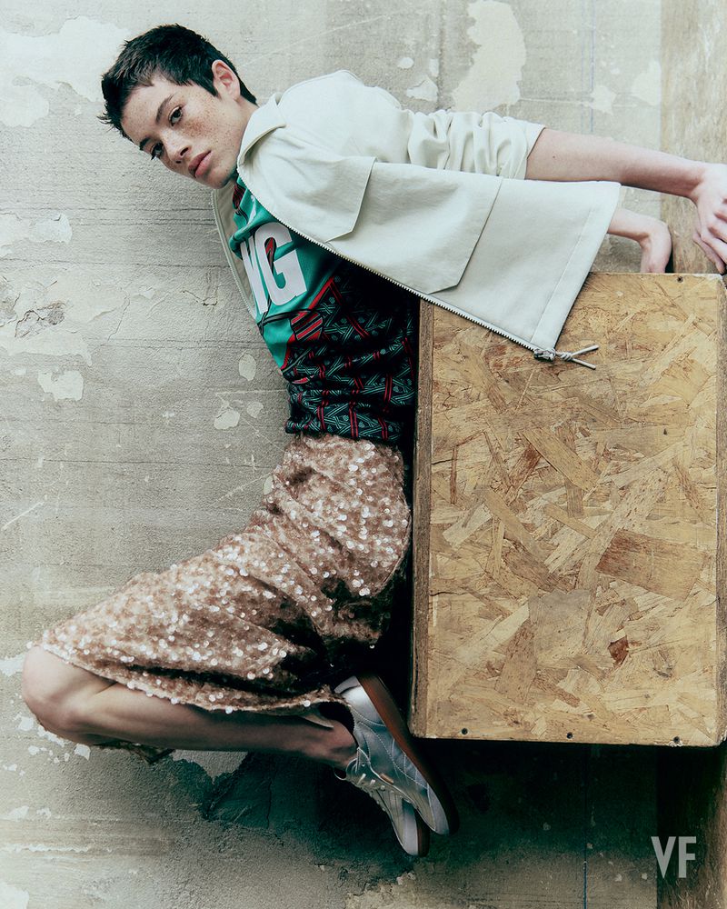 A non-binary person poses against a concrete wall, wearing a green graphic tee, jacket, sequined skirt, and metallic boots near a wood crate.