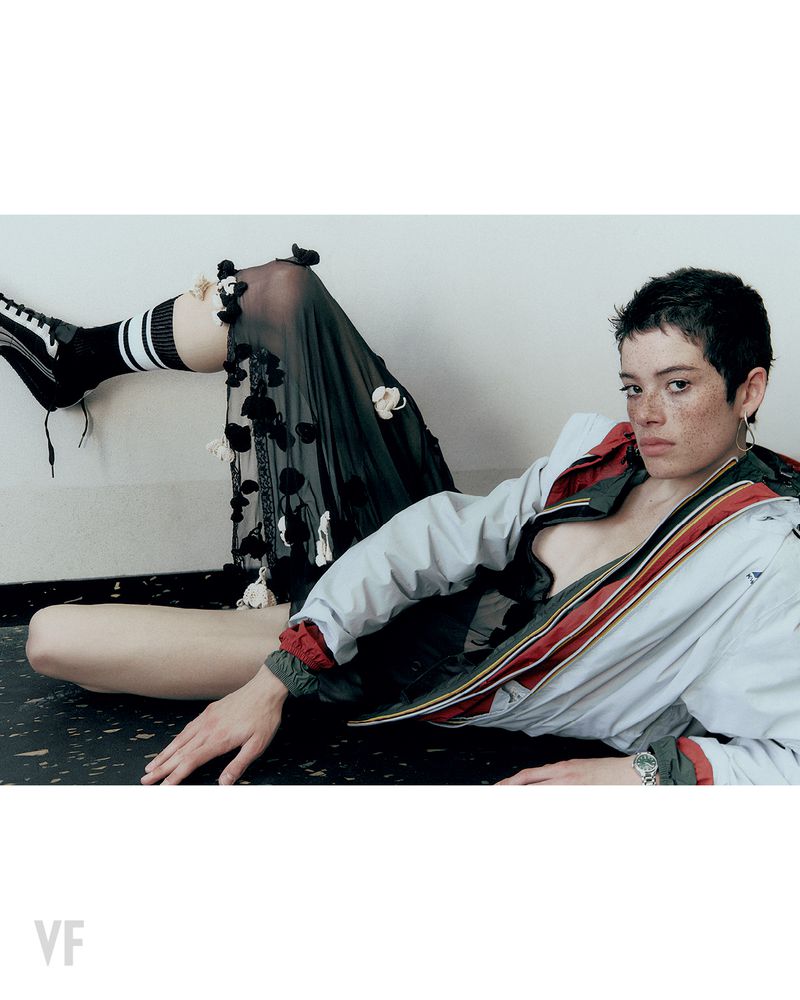 Model with short dark hair and freckles reclining in a windbreaker and sheer skirt for Vanity Fair Italia.