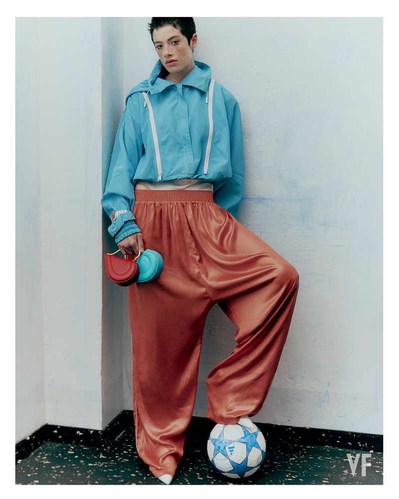 Model Dantè Kedde in a blue cropped windbreaker and terracotta satin trousers resting one foot on a soccer ball in a white room corner.