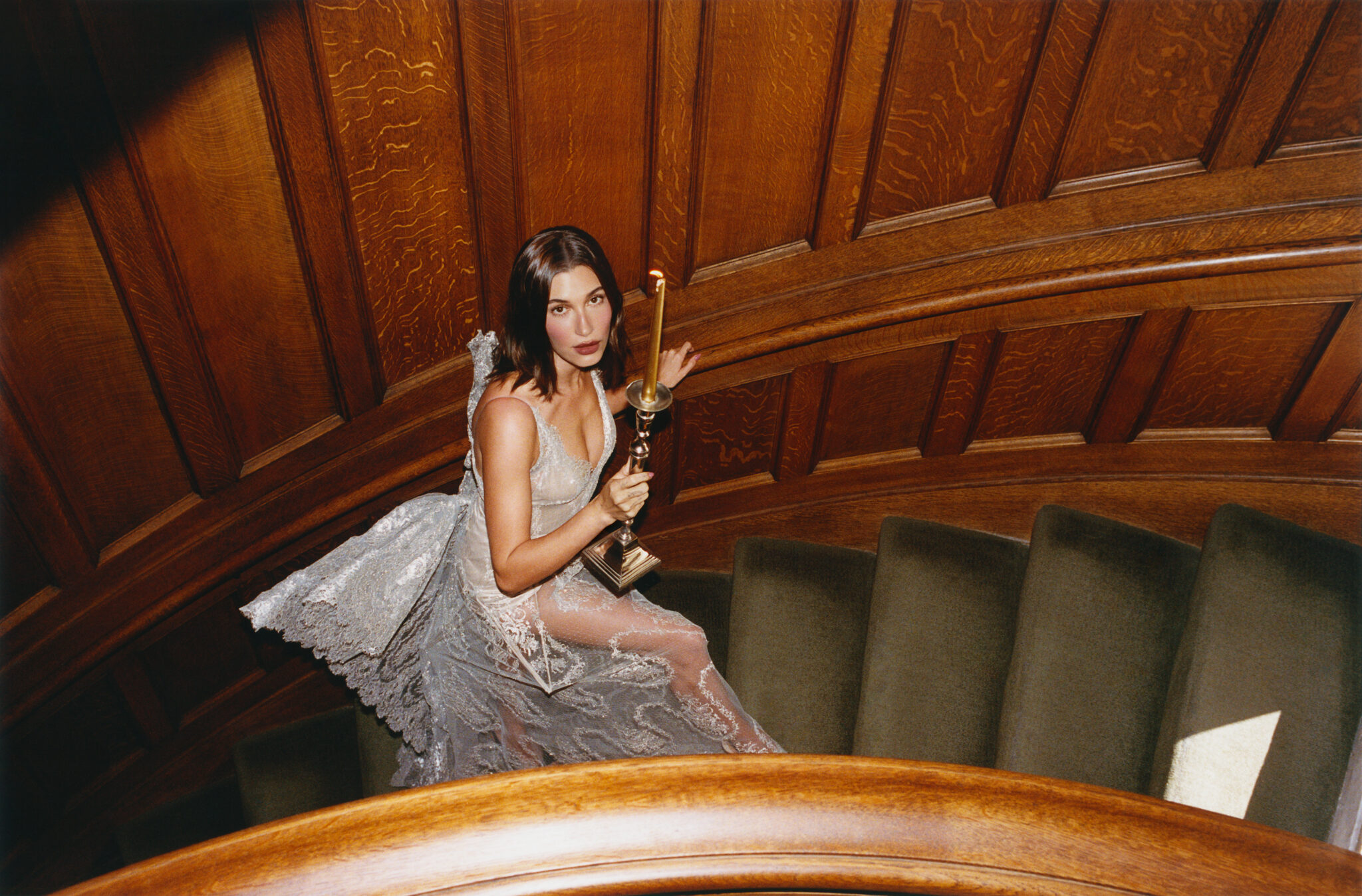Hailey Bieber in a sheer lace gown holds a candlestick while posing on a wood-paneled spiral staircase.