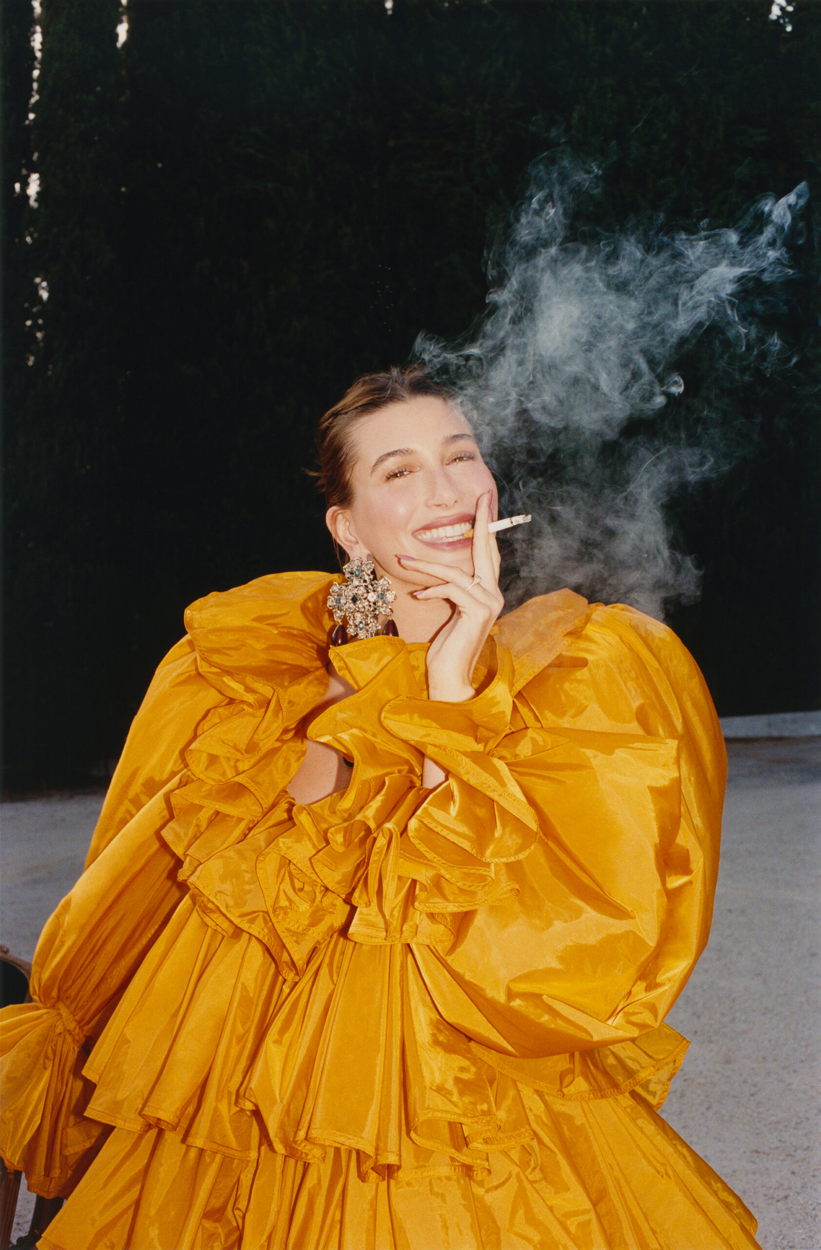 Hailey Bieber smiling in a voluminous yellow ruffled gown while holding a lit cigarette surrounded by smoke outdoors.