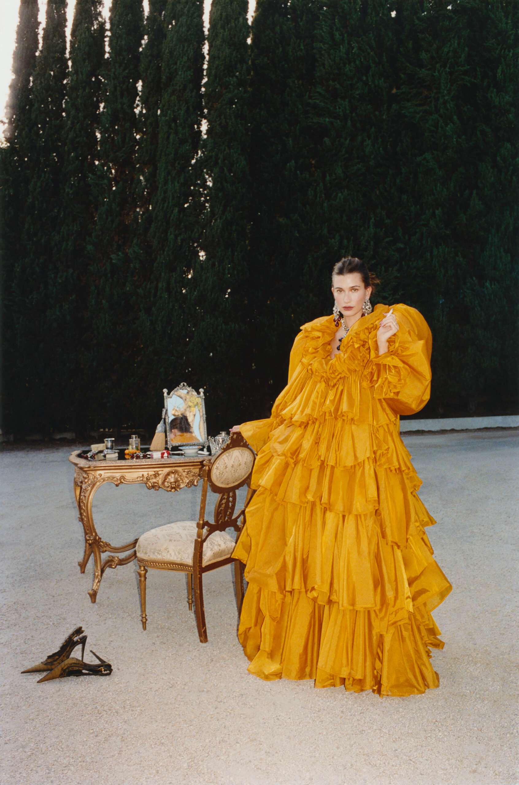 Hailey Bieber poses in a voluminous tiered yellow gown beside an ornate gold vanity table outdoors in front of tall cypress trees.