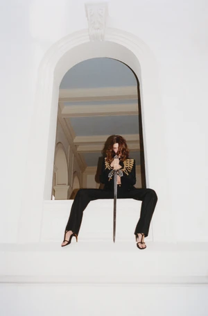 Model Hailey Bieber holding a sword while seated in an arched white window frame for Interview Magazine Spring 2026
