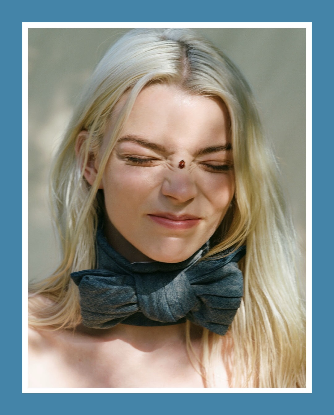 Close-up portrait of Anya Taylor-Joy squinting with a small beetle on her nose and wearing a grey fabric neck bow.