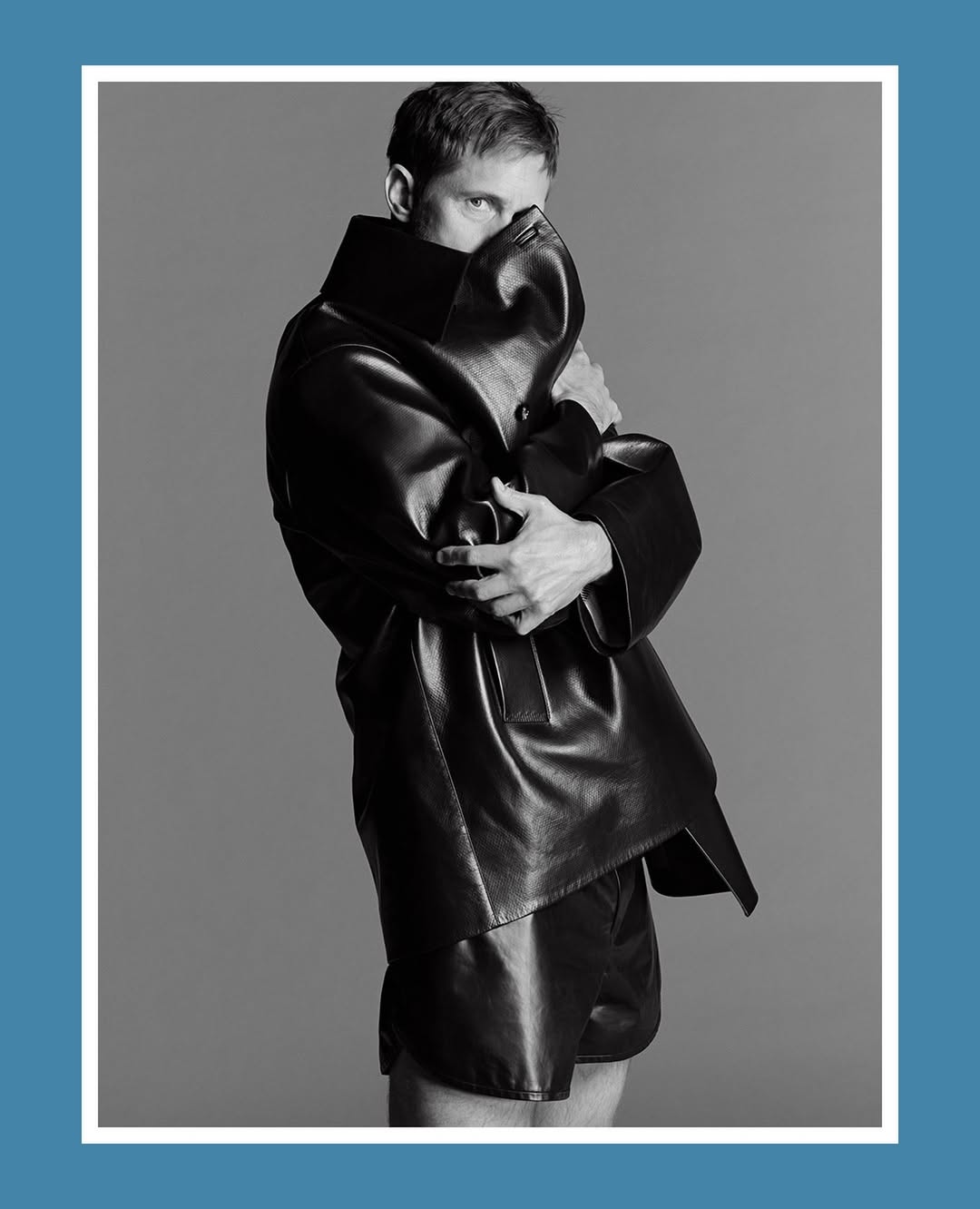 Alexander Skarsgård in an oversized black leather coat and shorts, partially covering his face against a grey studio backdrop.