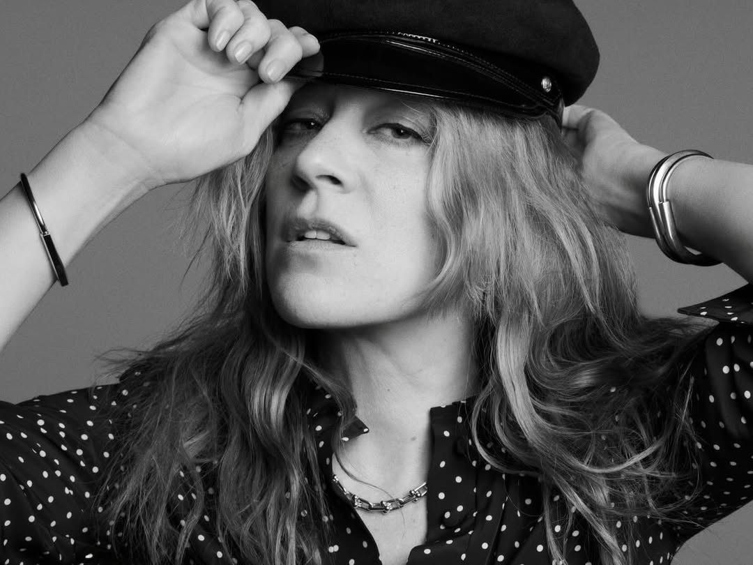 Black and white portrait of Chloë Sevigny wearing a black cap and polka dot blouse with hands raised to her head.