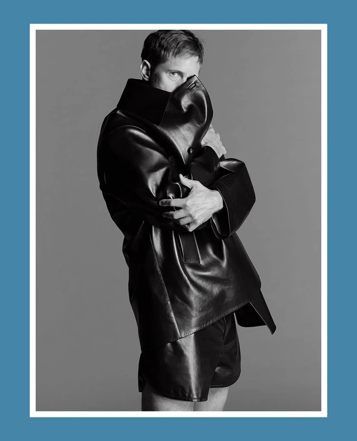 Alexander Skarsgård in an oversized black leather coat and shorts, partially covering his face against a grey studio backdrop.