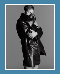 Alexander Skarsgård in an oversized black leather coat and shorts, partially covering his face against a grey studio backdrop.
