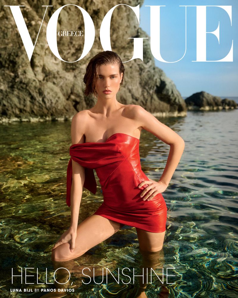 Model Luna Bijl wearing a red strapless mini dress standing in clear sea water by a rocky cliff for Vogue Greece.