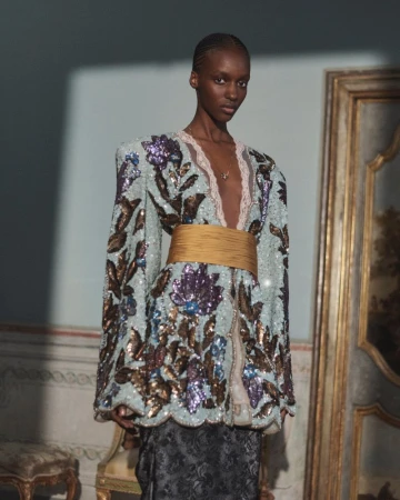 Model wearing a sequined mint floral jacket with a wide gold sash and deep V-neckline in an ornate classical interior.
