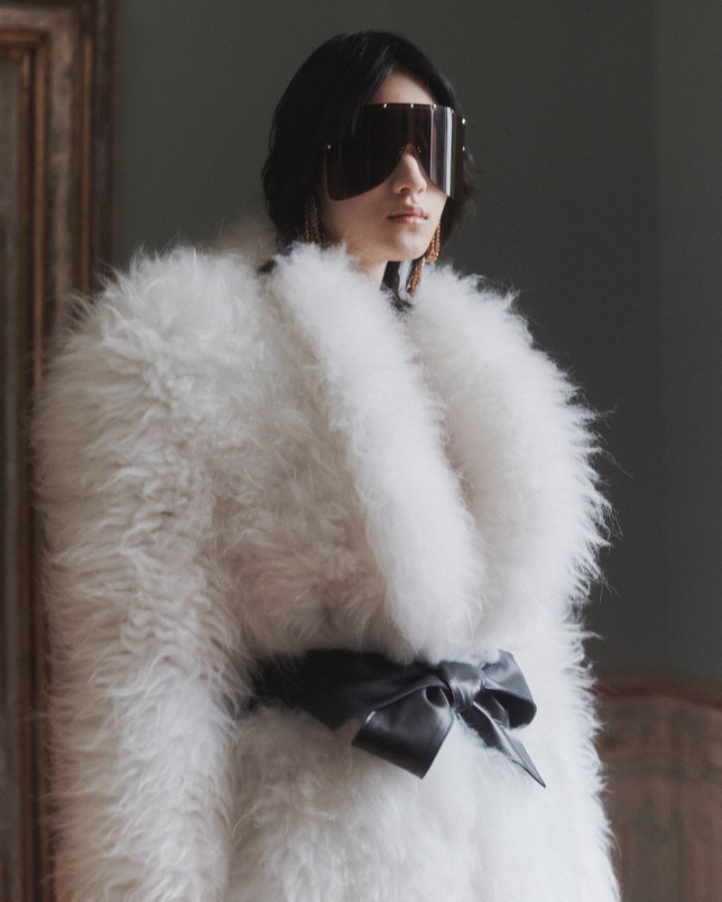 Model wearing a voluminous white shearling coat tied with a black bow belt and futuristic shield sunglasses in an interior setting.
