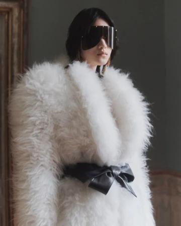 Model wearing a voluminous white shearling coat tied with a black bow belt and futuristic shield sunglasses in an interior setting.
