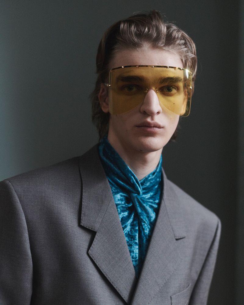 Model Saar Mansvelt Beck wearing yellow shield sunglasses, a grey blazer, and a teal crushed velvet ascot against a grey backdrop.