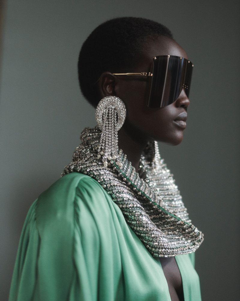 Model in profile wearing oversized shield sunglasses, crystal chandelier earrings, a heavy rhinestone cowl, and a green satin robe.
