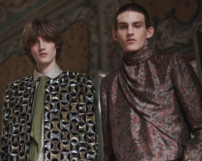Two male models posing in ornate high-fashion garments including a sequined geometric jacket and draped jacquard top.