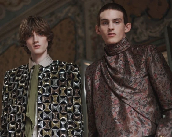 Two male models posing in ornate high-fashion garments including a sequined geometric jacket and draped jacquard top.
