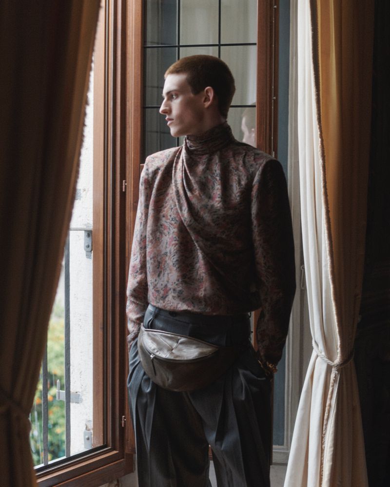 Saar Mansvelt Beck standing beside a tall window wearing a draped patterned high-neck top, pleated trousers, and a leather waist bag.