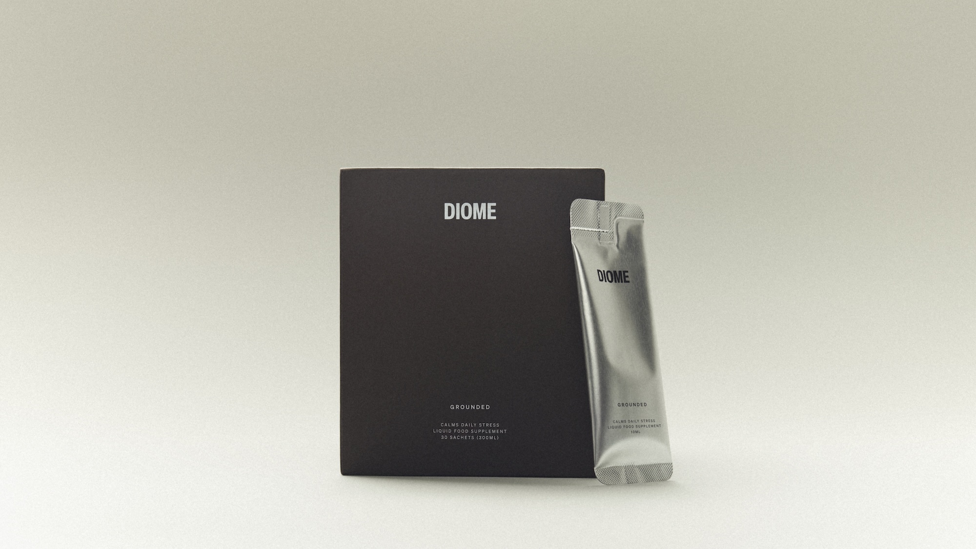 Diome Grounded supplement packaging box in matte dark brown standing beside a metallic silver liquid sachet on a seamless background.