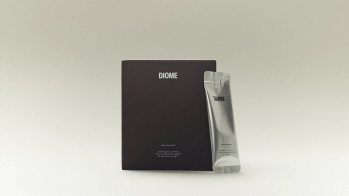 Diome Grounded supplement packaging box in matte dark brown standing beside a metallic silver liquid sachet on a seamless background.