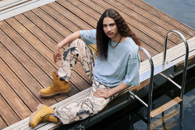A young adult male with long curly hair, wearing a light blue t-shirt and patterned pants, sits on a wooden dock with work boots.