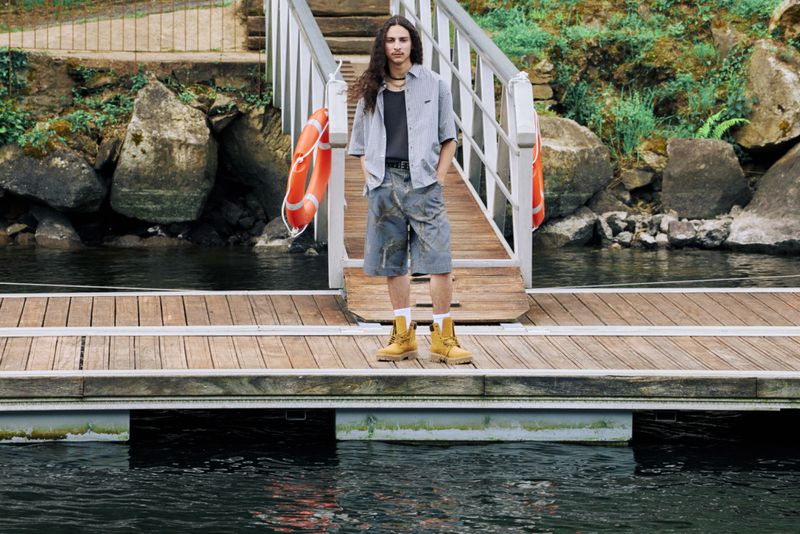 Man stands on a wooden dock in gray button-down shirt and patterned shorts, wearing tan boots, on a body of water by rocks.