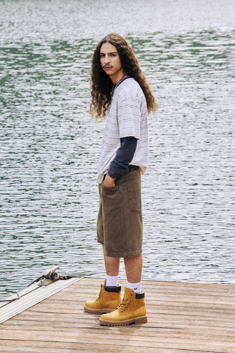 Man in casual wear stands on wooden dock with lake backdrop. Wearing tan work boots, olive shorts, striped tee. Long curly hair and mustache.