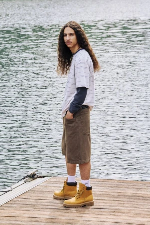 Man in casual wear stands on wooden dock with lake backdrop. Wearing tan work boots, olive shorts, striped tee. Long curly hair and mustache.