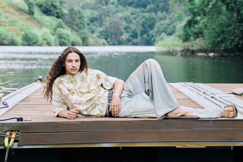Young adult male with long curly hair, wearing a patterned shirt and jeans, relaxing on a wooden dock by a lake with a forest backdrop.