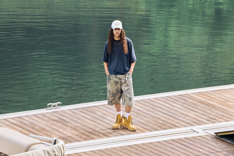 Model Rayan Mazuel standing on a wooden floating dock in an oversized blue shirt, patterned shorts, and wheat boots for Pull & Bear.