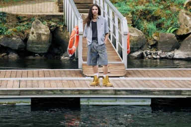 Model with long dark hair standing on a wooden dock wearing shorts, a striped button-down shirt, and yellow boots