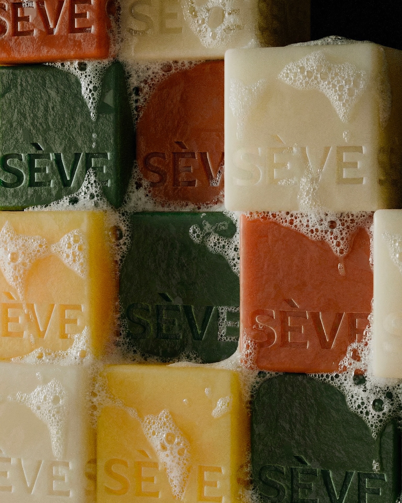 Stacked colorful bar soaps in green, terracotta, and cream covered in foamy soap bubbles and embossed with the Sève logo.