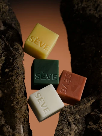 Four square embossed SÈVE soap cubes in cream, yellow, green, and terracotta framed between textured dark tree bark.