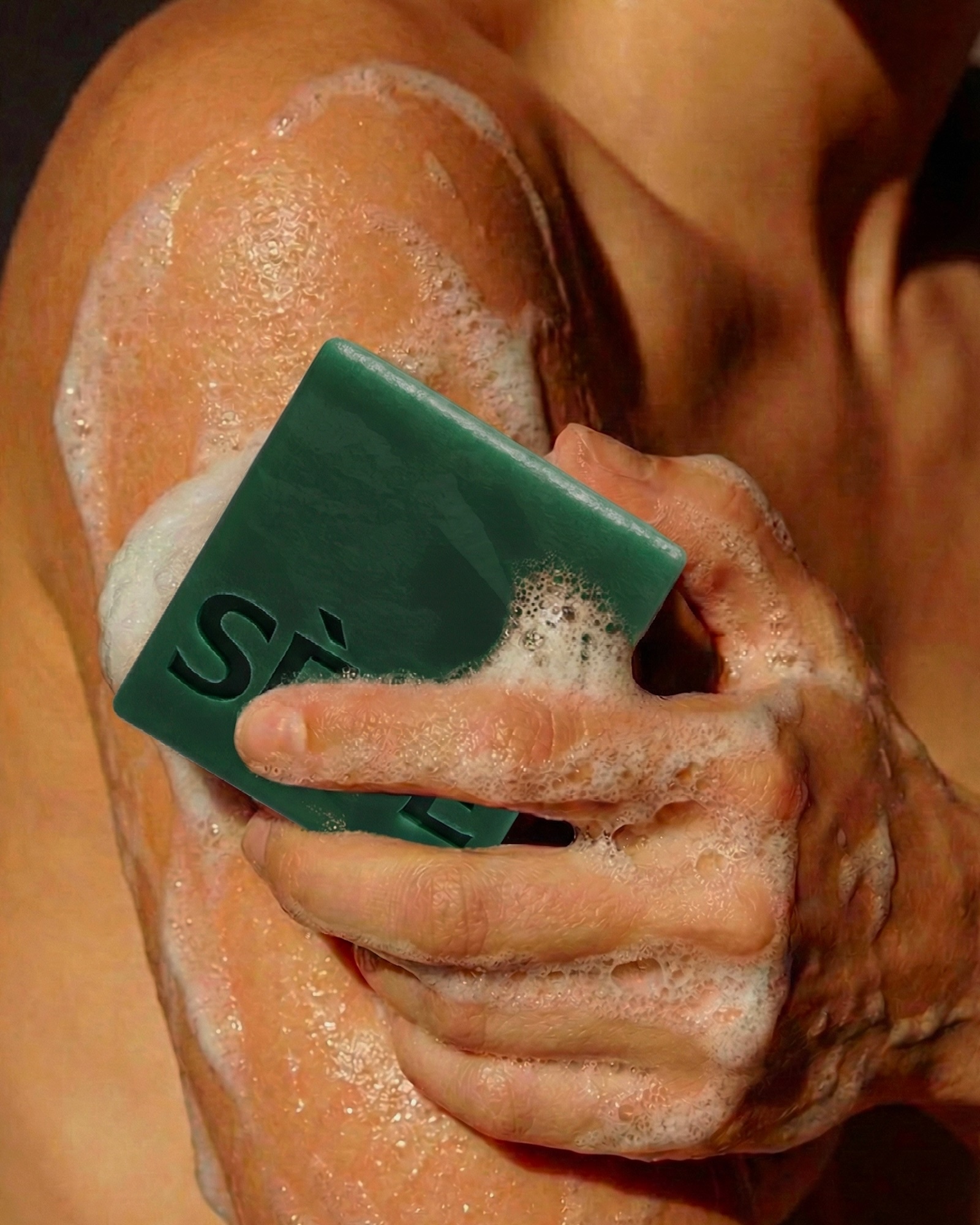 Close-up of a foamy hand rubbing a dark green bar of soap against a lathered shoulder with wet skin.