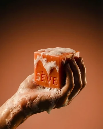 Soapy hand holding an embossed terracotta cubic bar of Sève soap covered in lather against a warm gradient background.