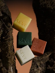 Four square embossed SÈVE soap cubes in cream, yellow, green, and terracotta framed between textured dark tree bark.