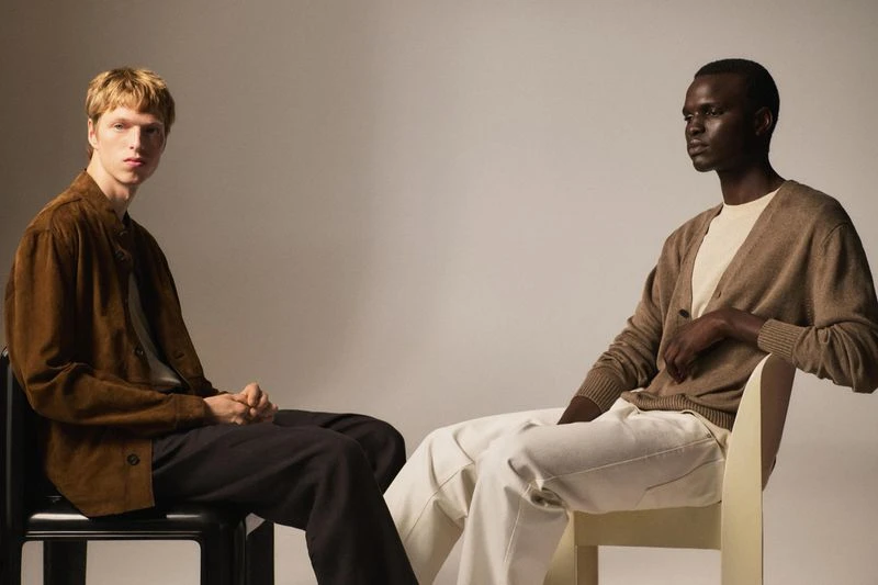 Two men sit in a photo studio, one in a brown jacket and the other in a cardigan, both in neutral pants, against a grey background.