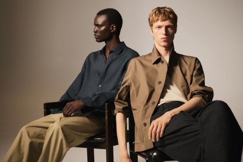 Two adult men sit indoors under studio lighting wearing casual fashion clothing in a portrait shot with a neutral background.