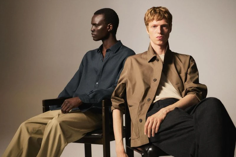 Two adult men sit indoors under studio lighting wearing casual fashion clothing in a portrait shot with a neutral background.